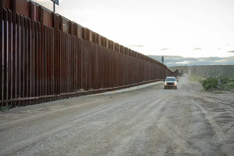 Border security patrol along a dirt road next to a large fence.